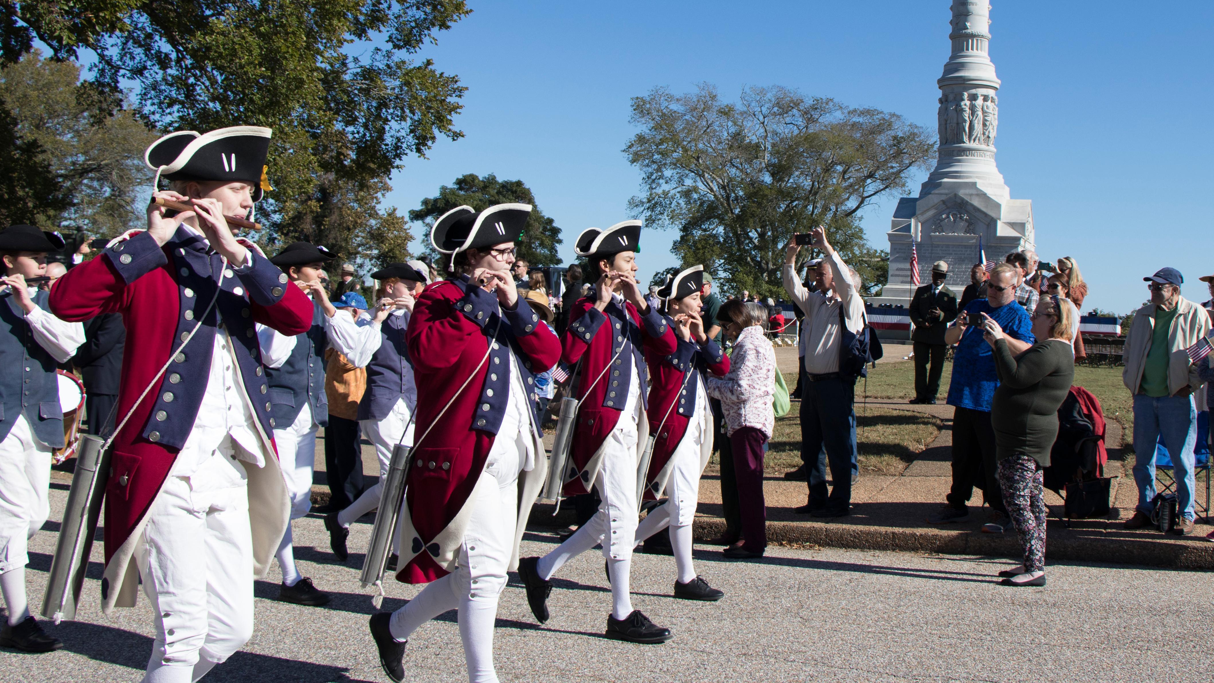 Fife and Drum wearing Red jackets play flutes in front of Victory Monument