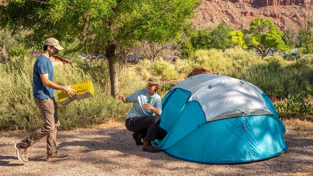 Two people set up camp next to a bright blue tent. Huge sandstone cliffs stand in the background.