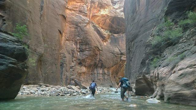 Two hikers wade through a river under towering walls.