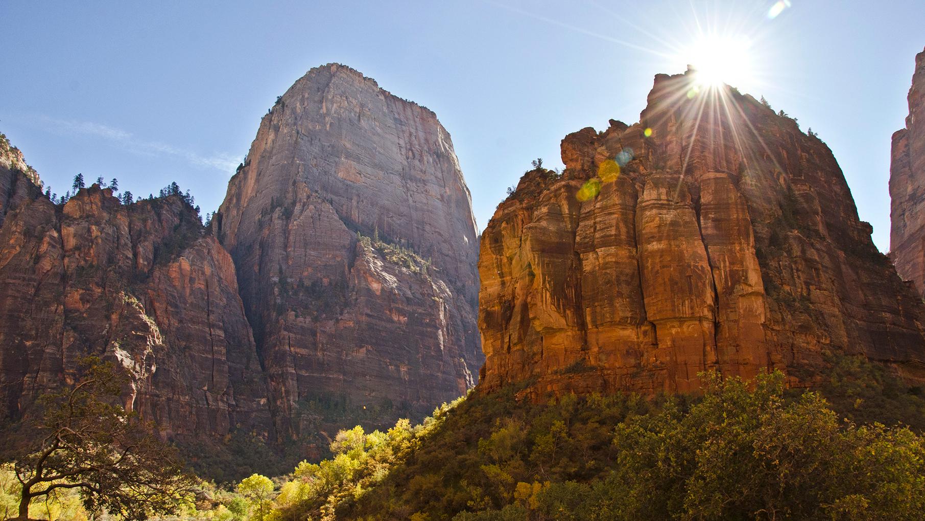 Sunshine peaks out from behind a red sandstone cliff. A tall mountain is in the background.