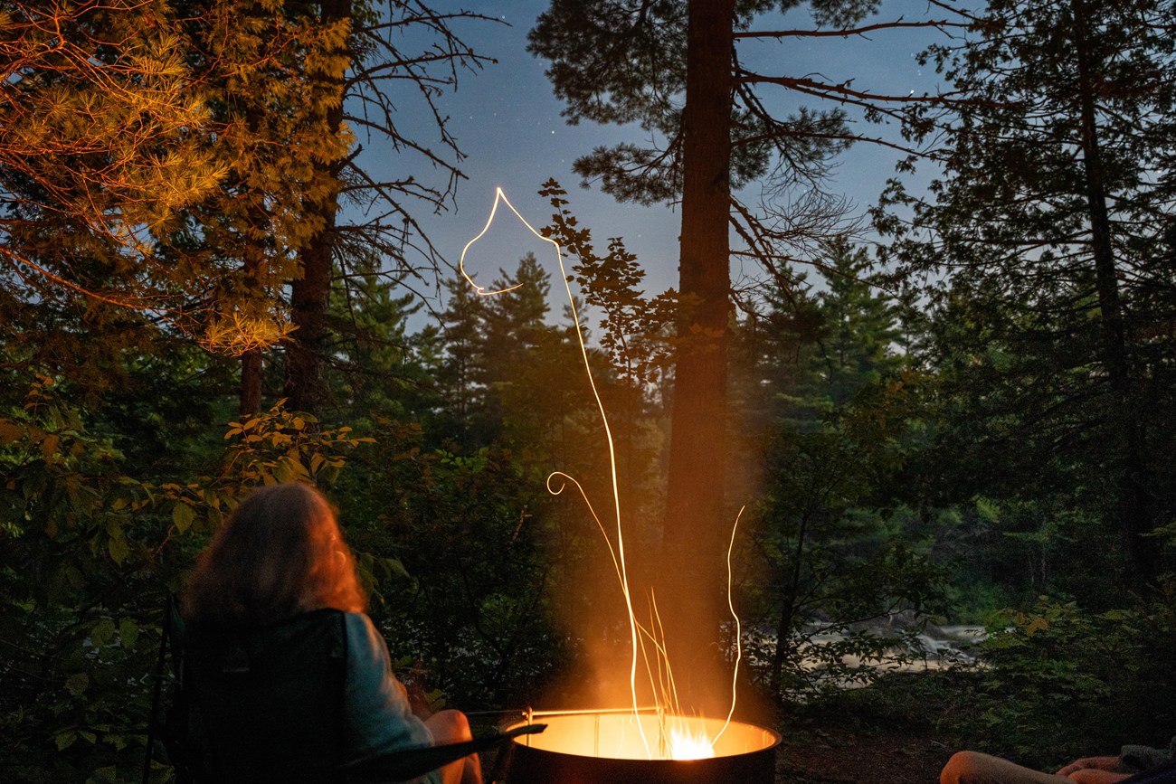 A bright orange fire coming from a fire ring lights up the person sitting in front of it. A river is visible in the background with rapid water flowing while the night sky above showcases stars.