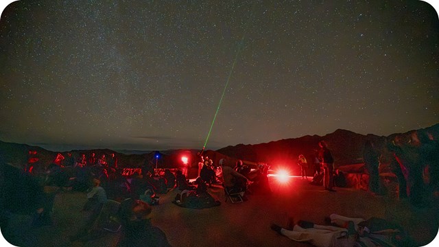 Beneath a star-filled sky, a crowd of people, illuminated by various lights, gather in the night. 