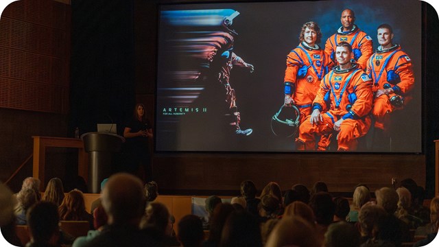 The photograph captures a speaker standing next to a large screen.