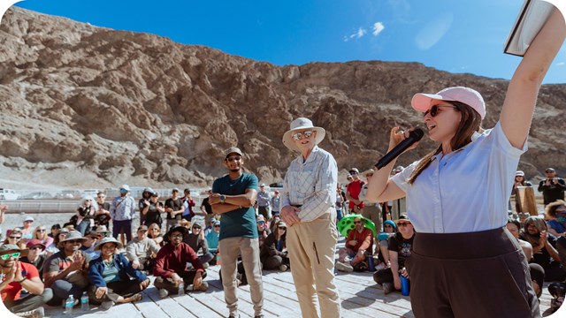 In this vibrant outdoor group scene, a woman speaks animatedly into a microphone.
