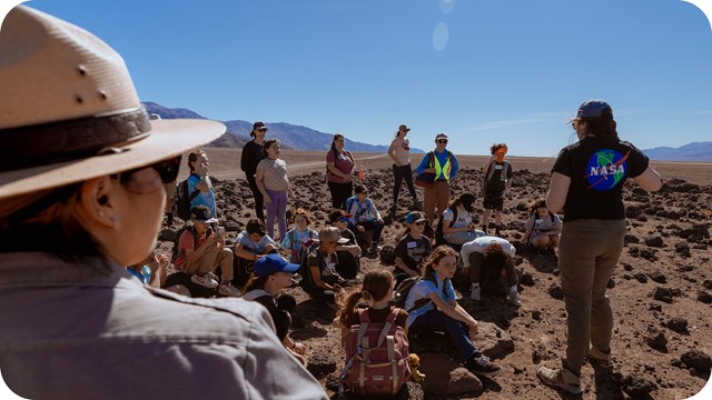 A group of people, mostly children and young adults, are gathered in an expansive, rocky desert.