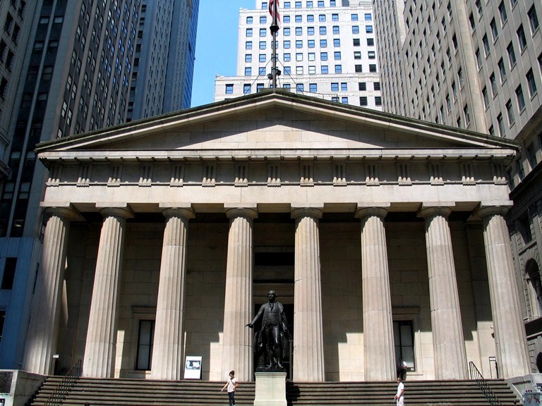 A neoclassical stone building with columns and a triangular roof, flanked by skyscrapers.