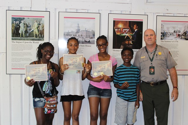 4 children and 1 park ranger standing together smiling while holding certificate and junior ranger badge