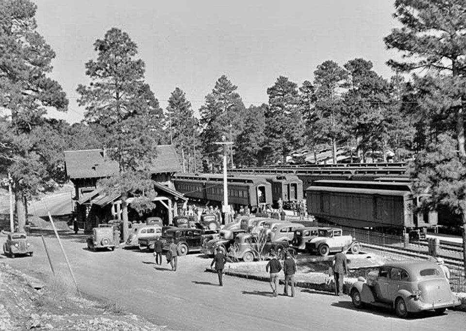 People, train, and cars next to railroad depot building, trees in background