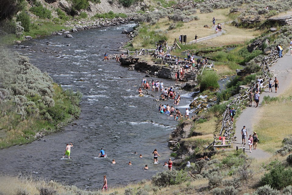 A river winds through the mountains. A large group of people are swimming in it and walking on a paved path beside it.