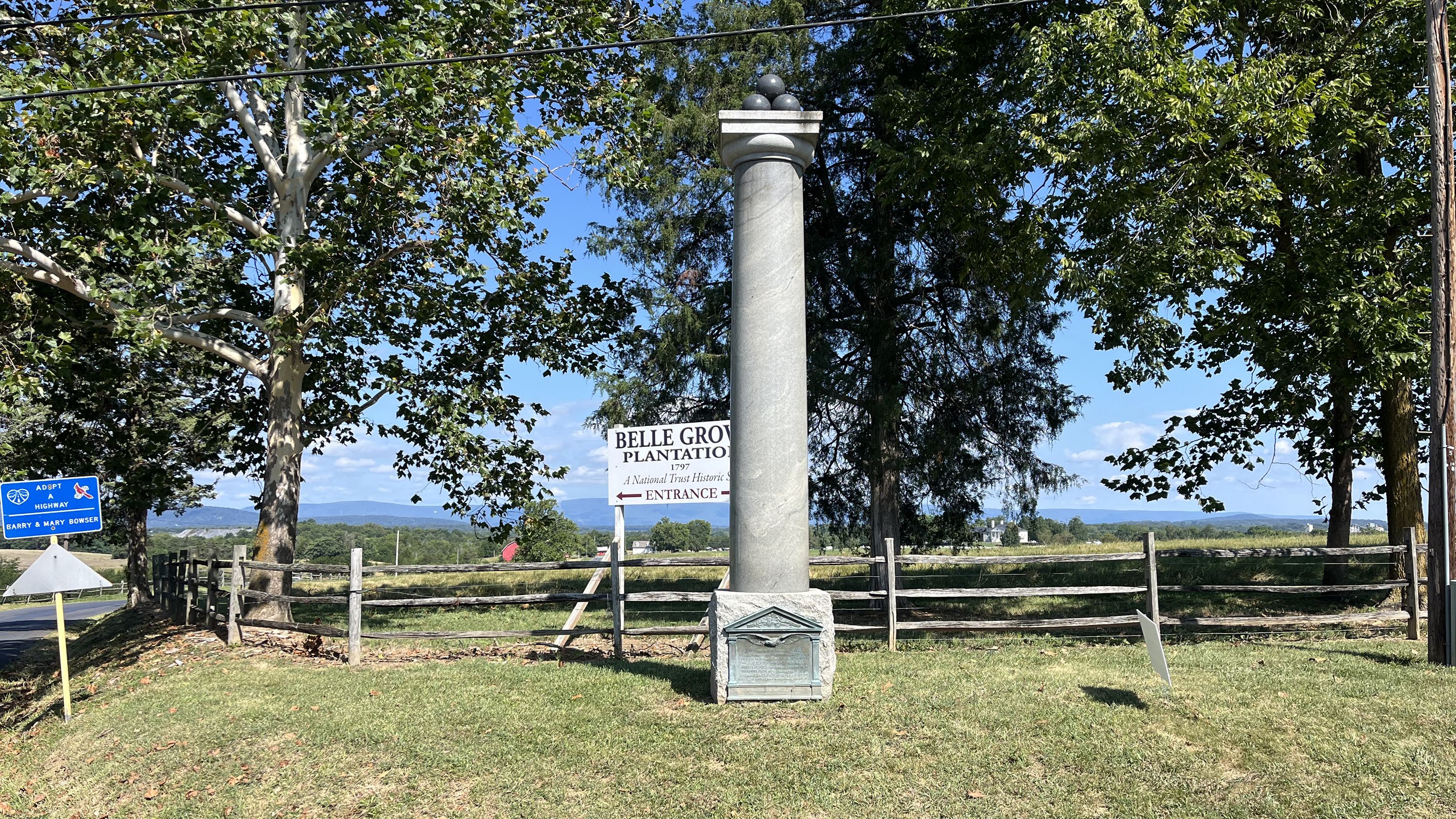 A tall granite column with cannonballs on top in front a wooden fence and a row of green trees.