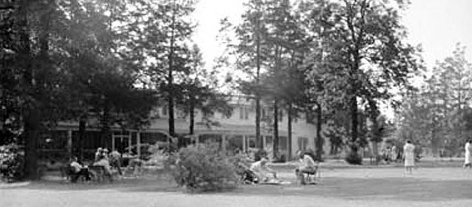 People gather in chairs and in the lawn in front of a two-story hotel behind a row of trees, black and white photo.