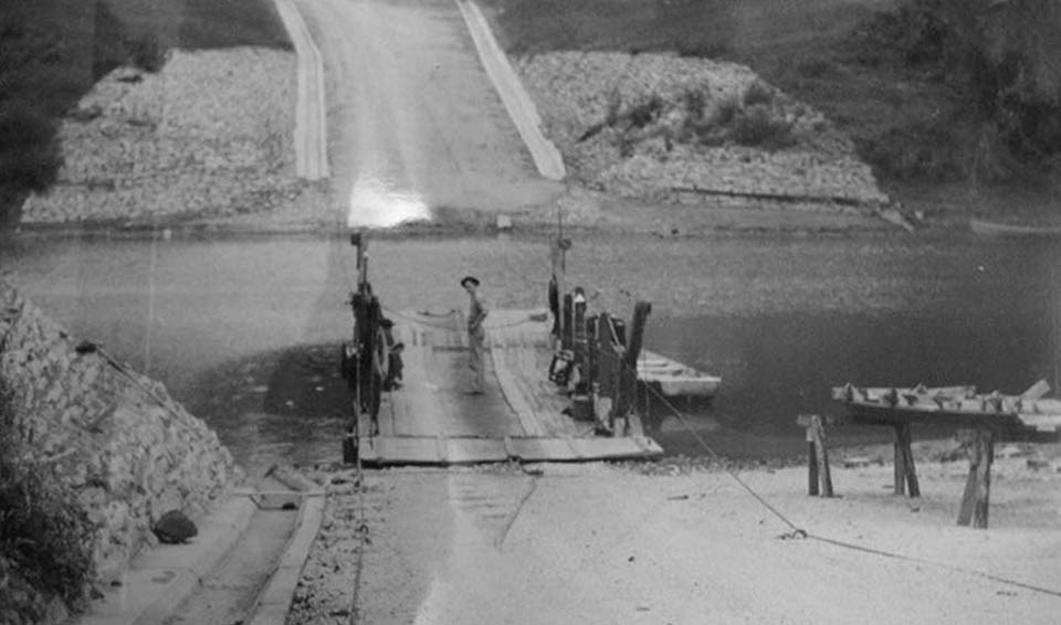 A person stands on wooden ferry at the end of a ramp, hands on hips. The road and concrete gutters continue on the other side of the water.