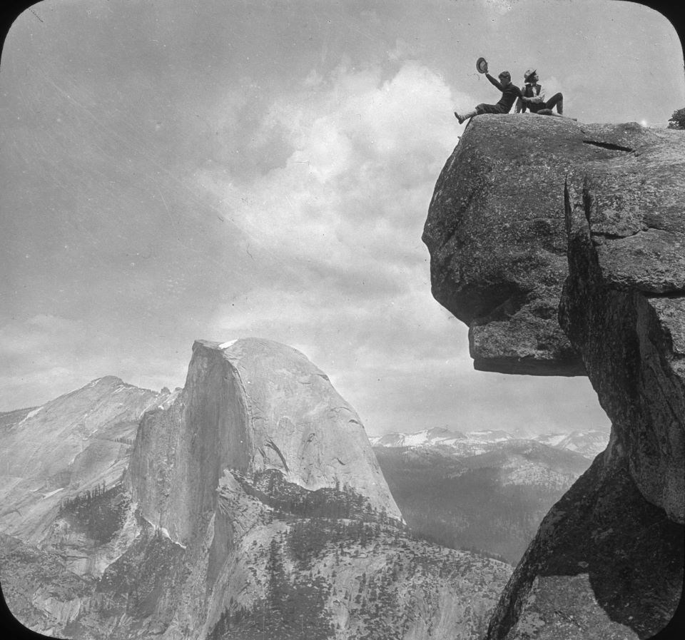 Black and white photo of two men on top of a rock overhang with Half Dome in the distance.