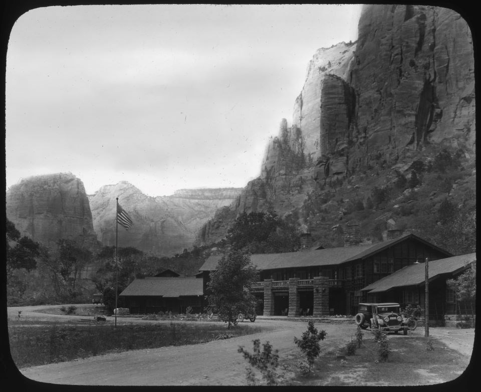 Black and white photo of a large wooden building with an old-fashioned car at the bottom of a cliff.