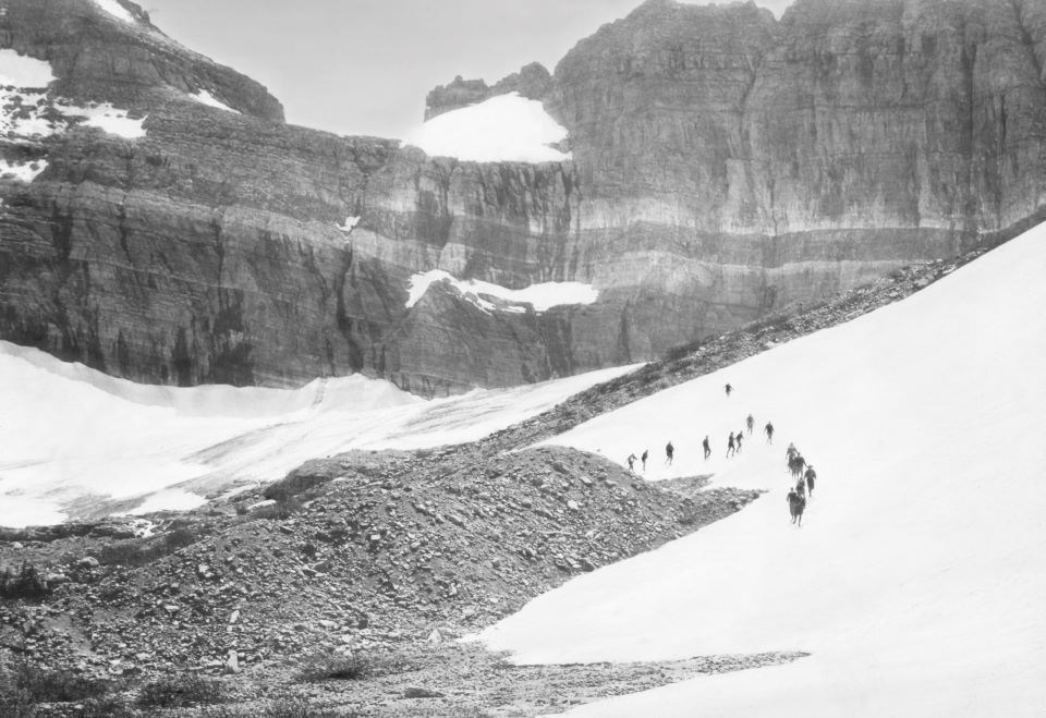 Black and white photo of a line of people walking in a snow filled valley.