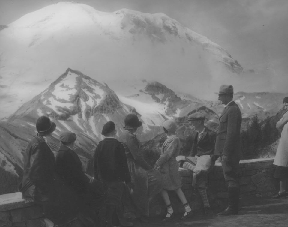 Black and white photo of people standing at a low wall looking at mountains in the distance.