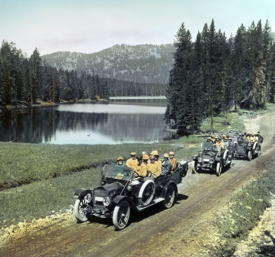 Black and white photo of three open top old-fashioned cars full of people on a dirt road with a lake, trees, and mountains.