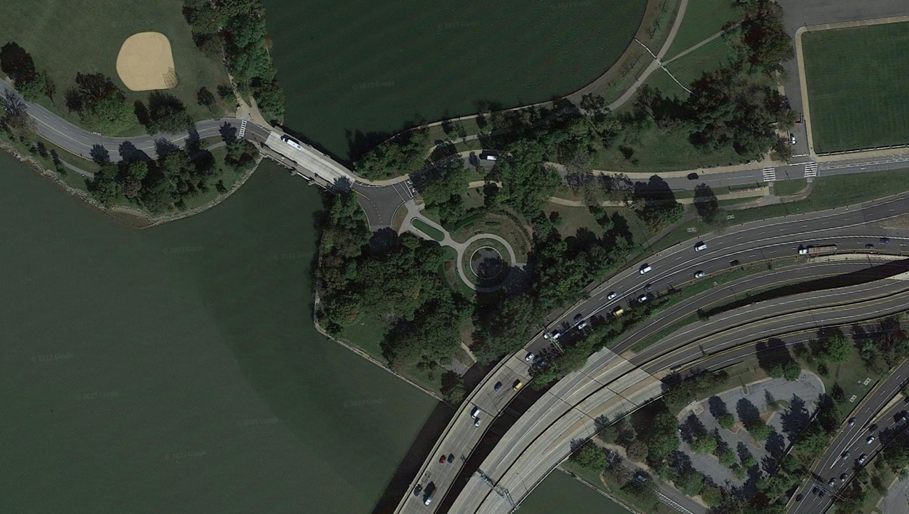 Aerial image of memorial landscape with round fountain and garden, thick canopy of trees, highway bridge over the river to the left, and cleared area to the right.