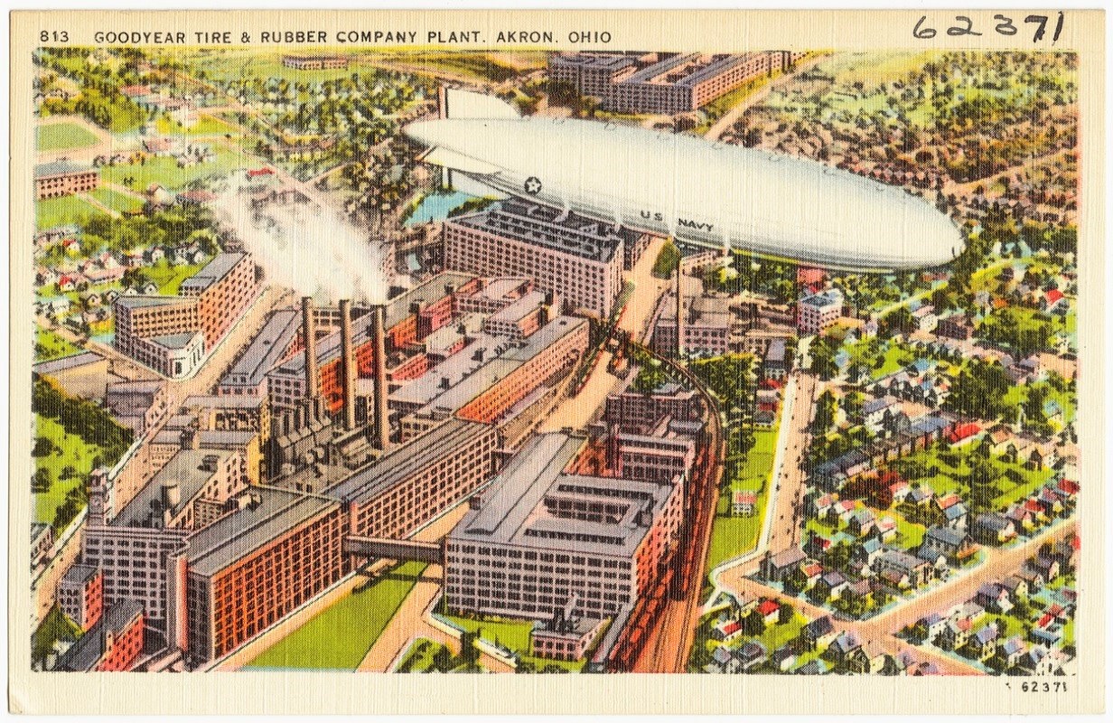 Color postcard illustration of bird’s eye view of blocky, multi-story factory buildings surrounded by a residential area. Three smokestacks spew smoke. Train tracks run alongside and through the complex. A white US Navy blimp flies overhead.