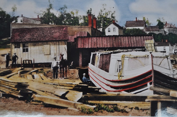 Colorized photo of canal boat, lumber, and workers in a boatyard along a canal.