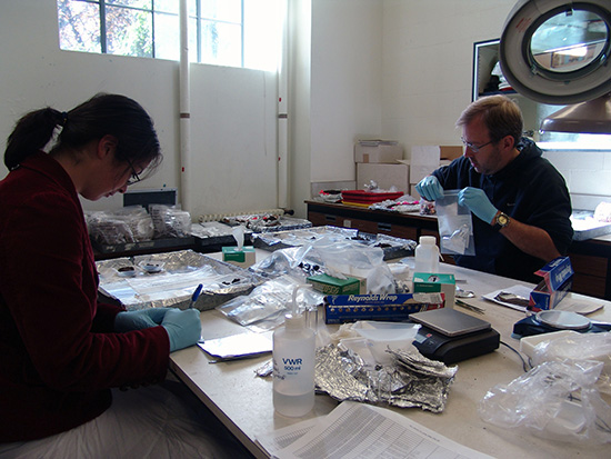 Two people sit at a table covered in artifacts and cataloging equipment Two people sit at a table covered in artifacts and cataloging equipment