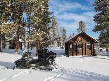 Small, brown wooden building covered in snow, standing amongst conifer trees with a snowmobile in the foreground.