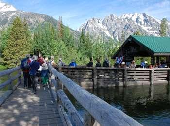 Visitors lined up to board a shuttle boat on the east side of Jenny Lake. Some standing on bridge over Cottonwood Creek, some standing on dock. Dock is partly covered by roof, but is open-air.