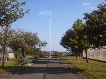 A thirty-foot tall flag pole stands at the end of a wide walking path, lined with grass and trees.