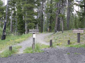 Trail leading off from a gravel parking area, with a wooden sign warning that it is closed until July 1.