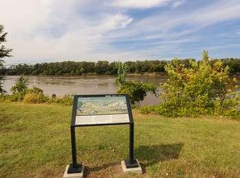 An interpretive wayside sign stands in a grass clearing with the large Missouri River flowing behind it