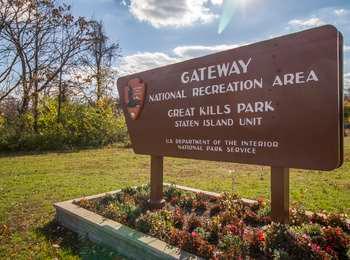 A brown Gateway National Recreation Area road sign with the traditional NPS arrowhead logo, sits amid a raised bed of begonias.