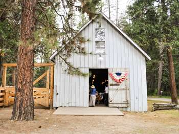 One door of a grey barn is open. Rows of lights hang from the ceiling and people can be seen sitting inside. Trees surround the area.
