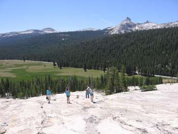 Several people stand on top of a granite dome which gives views of a large, open meadow surrounded by pine trees. Granite peaks rise above the treeline.