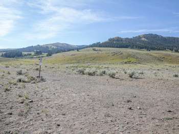 A bare ground path leads across sage-covered hills toward the distant tree-covered mountains.