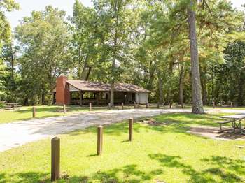 Wooden open air picnic pavilion. A wooden picnic table and a cement road stretching across green grass in the foreground. Green trees in the background.