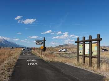 Looking north along the paved multi-use pathway just south of Gros Ventre Junction. Vehicles on the road and Blacktail Butte to the east and the Teton Range to the west.