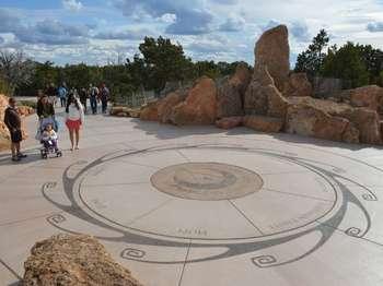 Visitors with a stroller walk next to a large bronze circular plaque set in the center of an open, concrete court area. The bronze plaque is decorated with an image of the canyon and a traditional southwestern petroglyph-style hand in the center. Surrounding the plaque is a large sun motif etched into the cement, along with the names of eleven local Native American tribes. Large, looming pieces of white-tan limestone encircle the court.