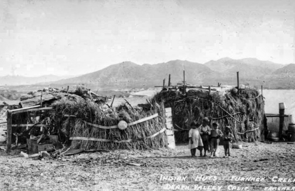 Children stand in front of straw huts with mountains in background.