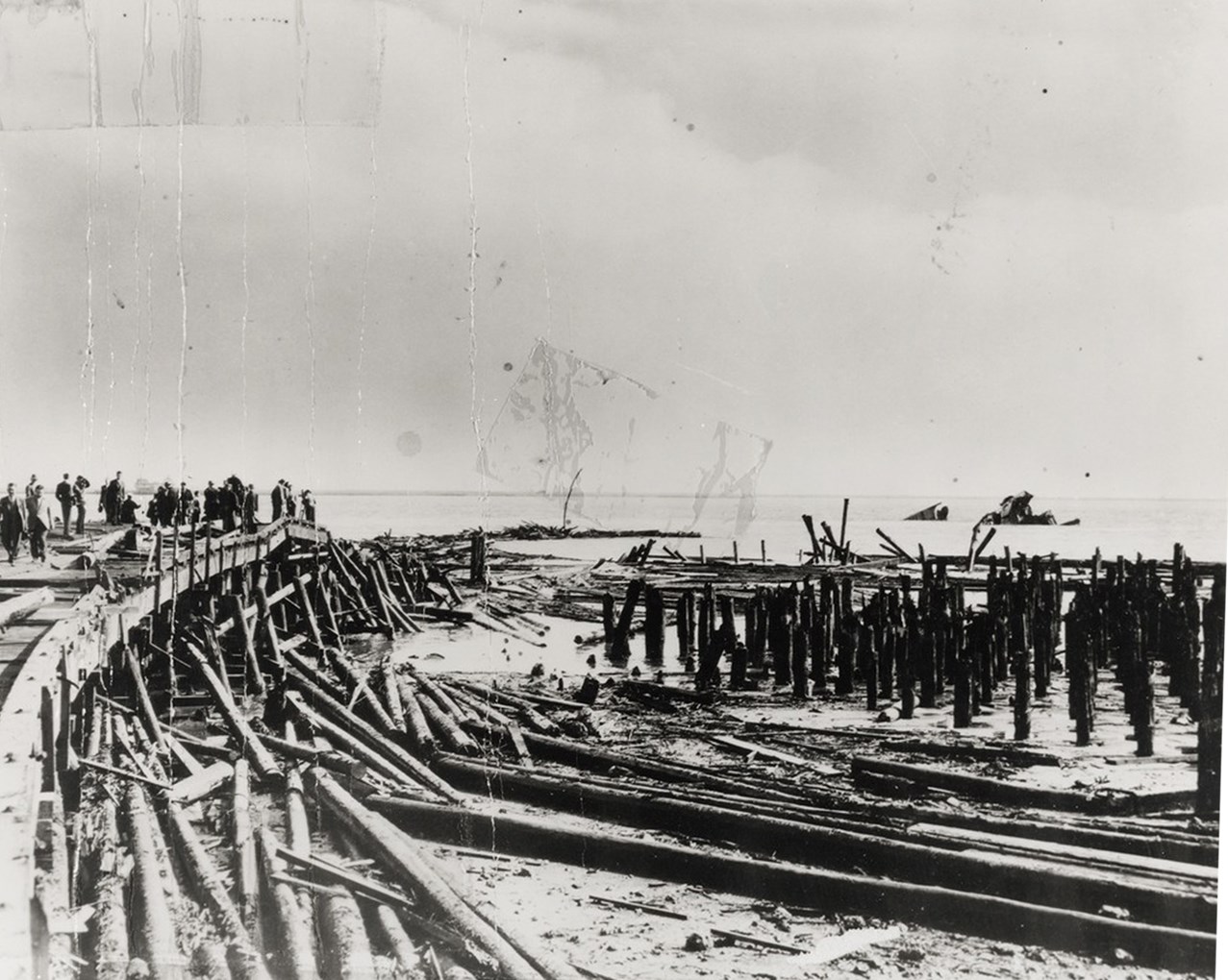 Historic black and white photo of the damaged pier. Debris is everywhere. Bay water is seen.