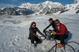 three people crouched near each other on a glacier under blue skies