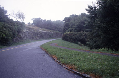 A road, framed by a rock retaining wall, winds through a hilly and partially-forested landscape. 