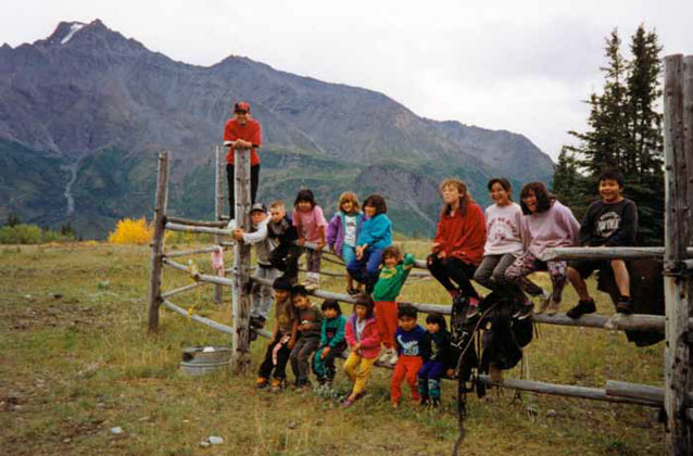 sixteen kids of various ages posing for a photo on a split rail fence in a mountainous landscape