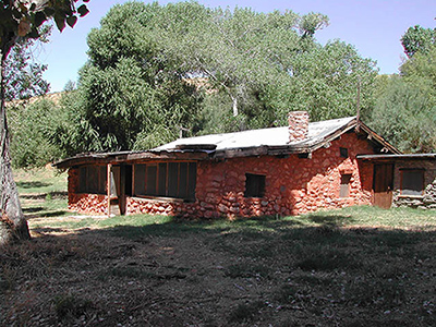 A single-story house of rough stone construction stands in the shade of leafy cottonwood trees.