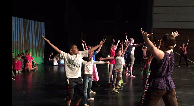 Students stand on stage raising their arms high above their heads.