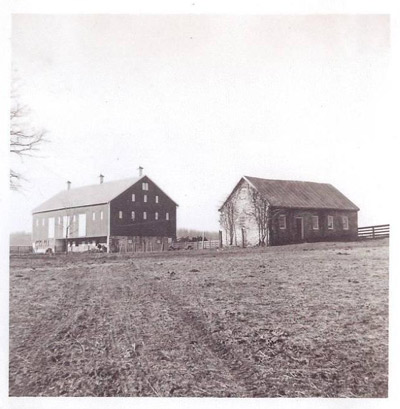 Two rectilinear farm buildings stand between an open, empty field and an open sky.