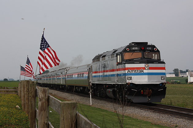 A silver Amtrak train with red, white, and blue stripes moves past a line of American flags. 