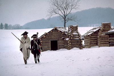 Two Revolutionary War reenactors walk in the snow with a log cabin and trees behind them