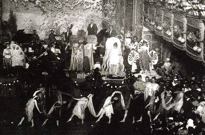 A line of dancers in costumes in front of a stage with a band and performers