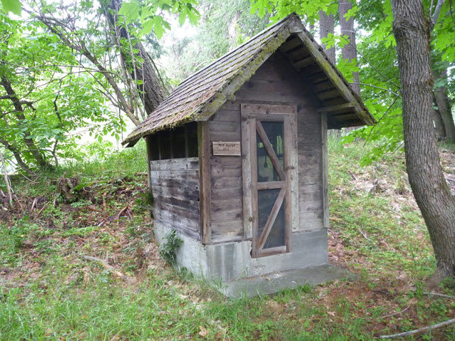A small, square wooden building with a peaked roof and one door in a wooded area