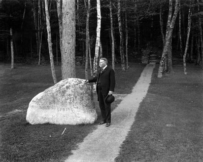 A man in a dark suit, holding a bowler hat, stands with one hand on a large boulder. 